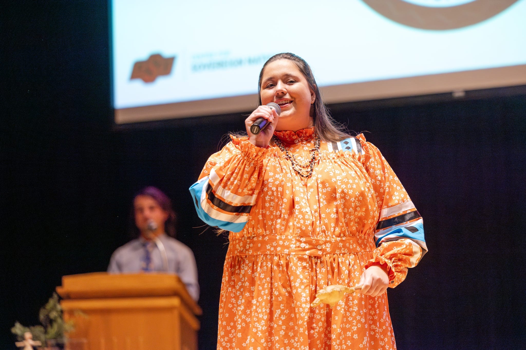 Photo of Miss American Indian OSU 2026-27 Kellon Collington in a traditional orange tear dress holding a microphone and a turtle shaped shaker