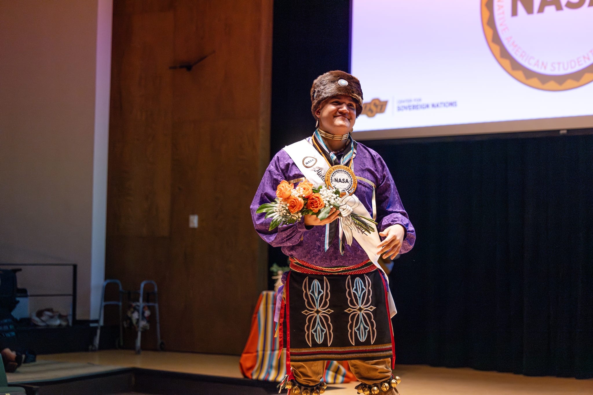 Photo of Mister American Indian OSU 2026-27 Elijah Grass in a purple ribbon shirt wearing a pelt hat holding flowers