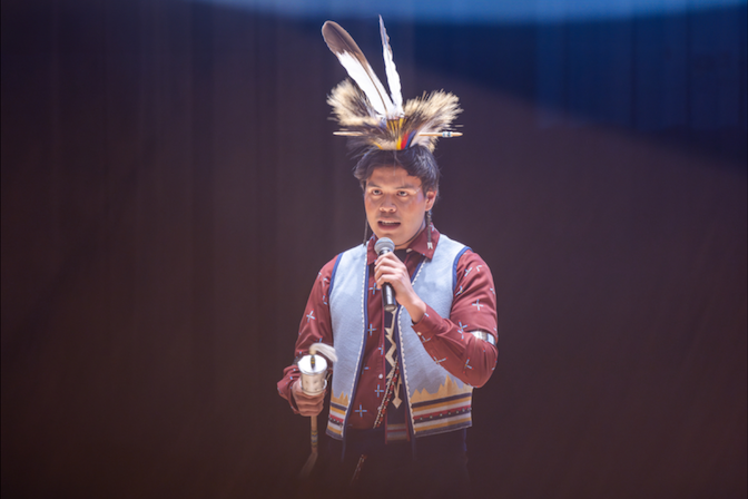 2025–2026 Mister American Indian OSU, Jaden Waters, a Kiowa citizen and OSU sophomore majoring in History (Pre-Law), speaks on stage while wearing traditional clothing—a ribbon shirt, vest, and roach—and holding a gourd rattle.