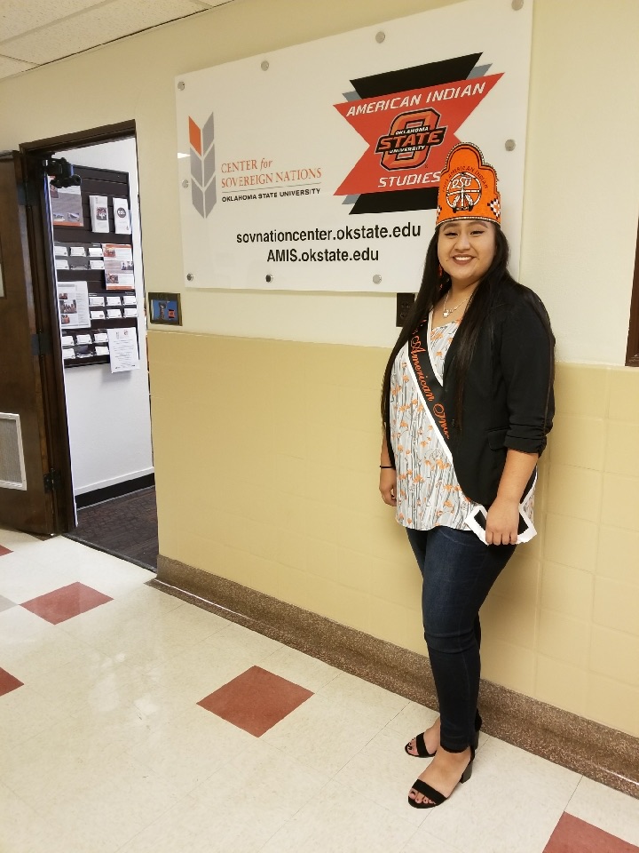 Miss American Indian OSU Cydney standing in front of the Center for Sovereign Nations American Indian Studies sign, smiling, wearing jeans, a sweater, and the MAIOSU crown and sash.
