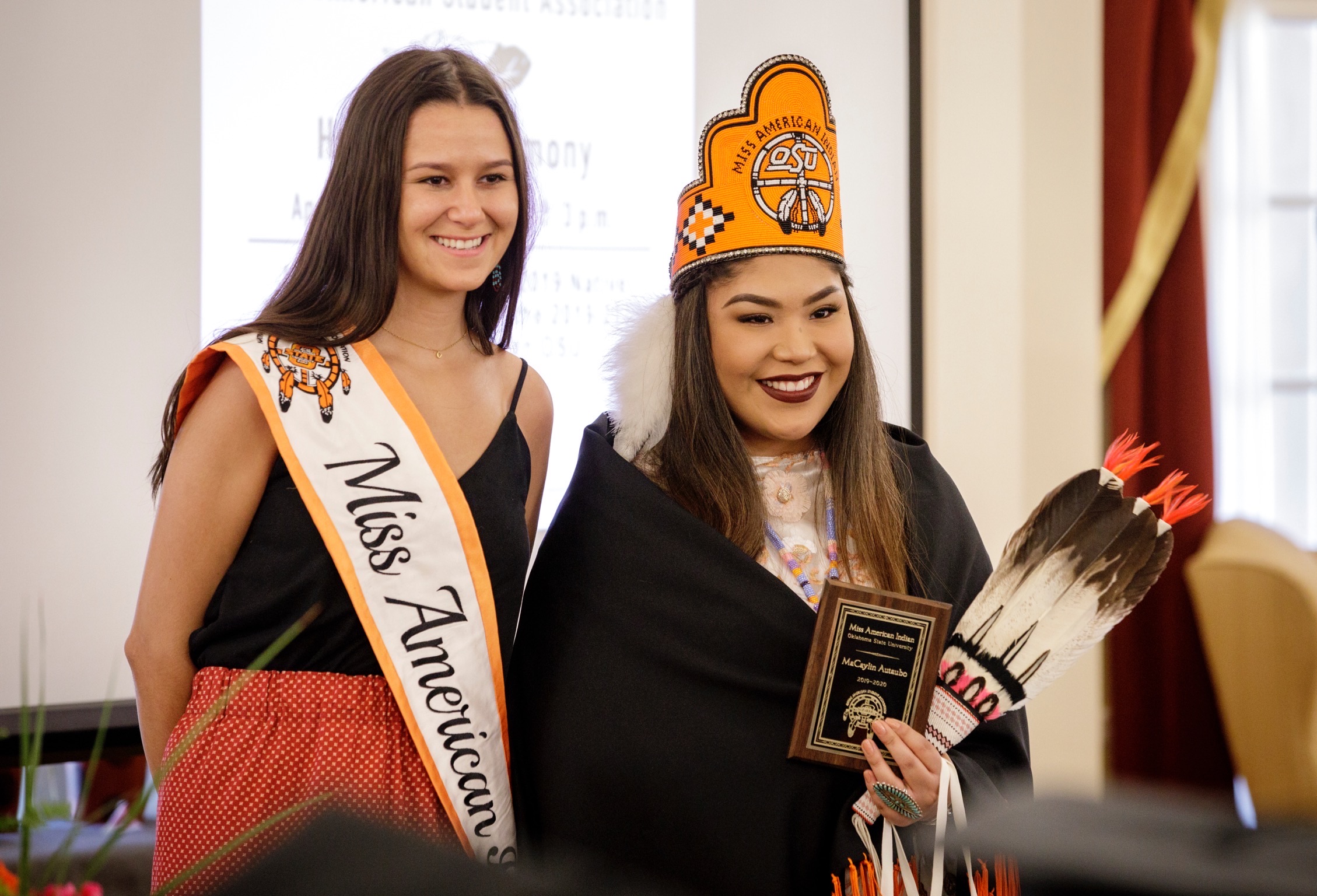 Miss American Indian OSU MaCaylin pictured with a former MAIOSU titleholder Cathrine. The former titleholder wears a ribbon skirt. Both women are smiling and looking at the camera, wearing their MAIOSU sashes, while the newly crowned Miss wears the MAIOSU crown and shawl and holds a traditional women’s feathered fan.