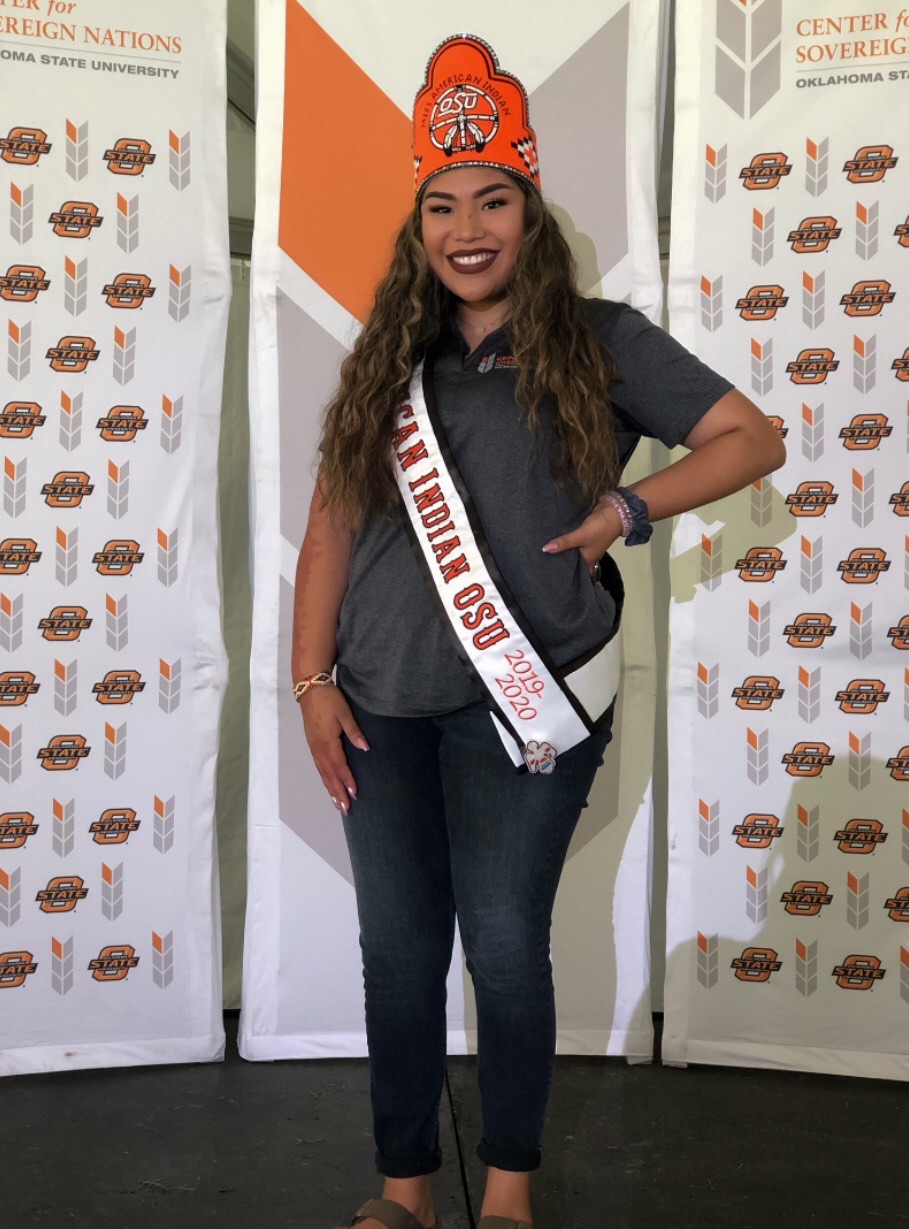 Miss American Indian OSU MaCaylin standing in front of Center for Sovereign Nations popup signs, smiling at the camera. She is wearing her MAIOSU crown and sash, a Center for Sovereign Nations polo, and jeans.