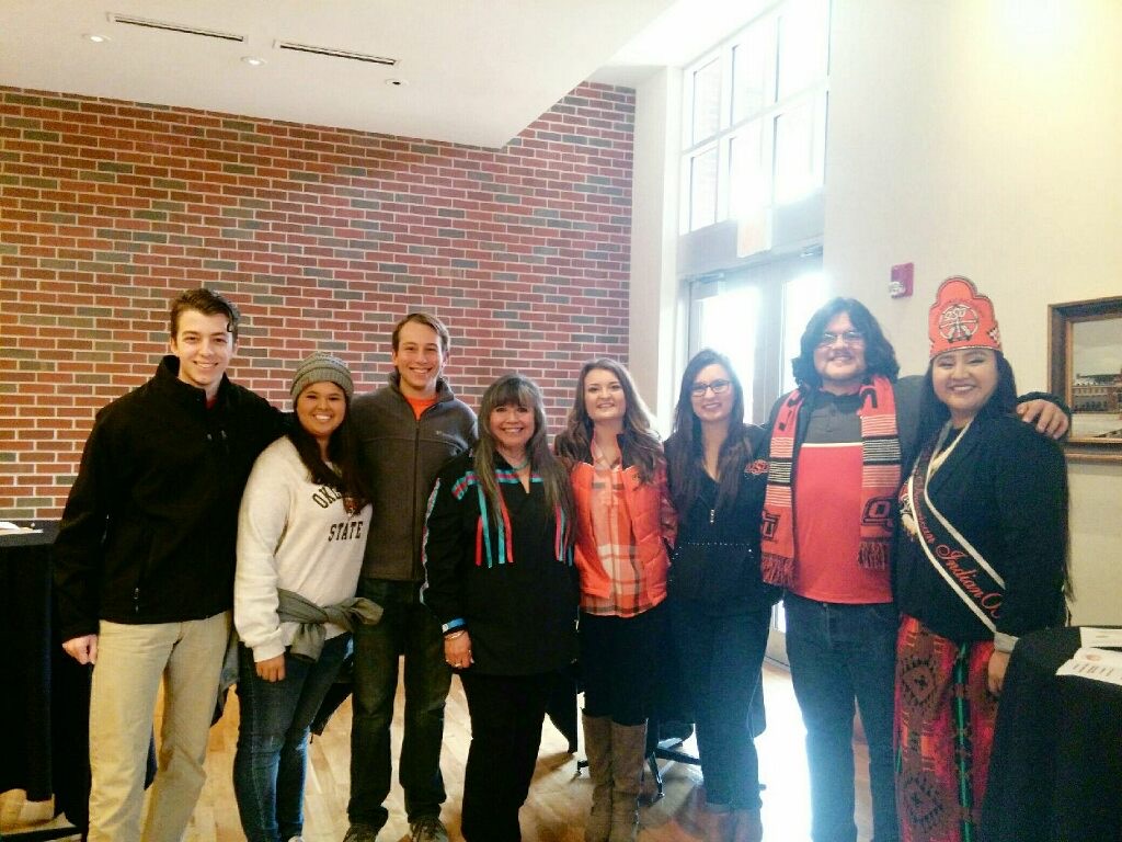 Miss and Mister American Indian OSU Cydney posing in a group photo with other NASA executive members, smiling at the camera. Both are wearing their sashes, and Miss Americ