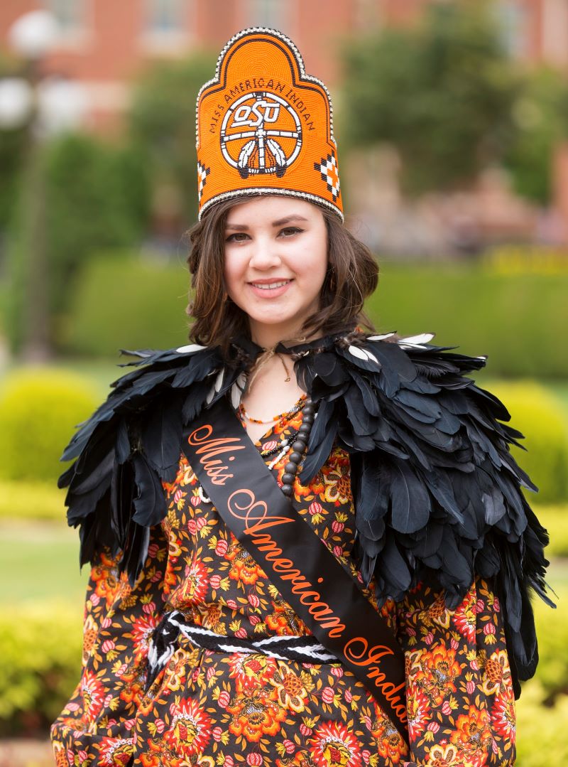 Miss American Indian OSU Meg, wearing the MAIOSU crown and sash along with a Cherokee Tear Dress and Cherokee feather cape, standing at the OSU Gardens.