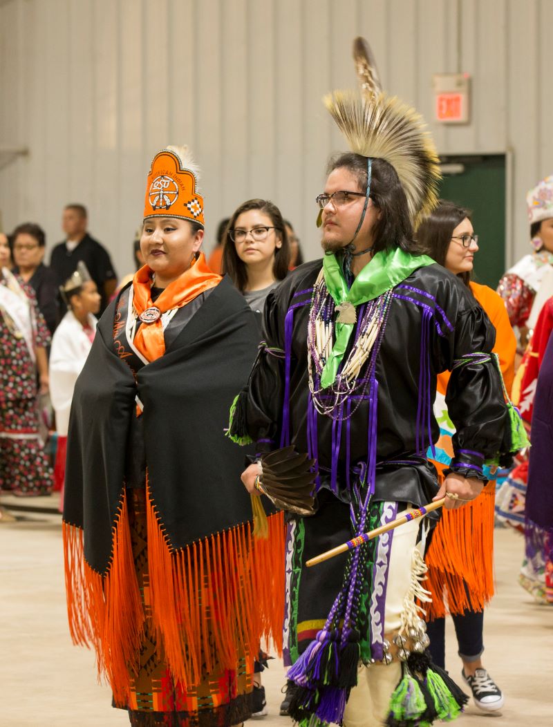 Miss American Indian OSU Cydnee and Mister American Indian OSU Che standing together, wearing MAIOSU crowns, sashes, and shawls, dancing together during Grand Entry at the OSU Powwow.
