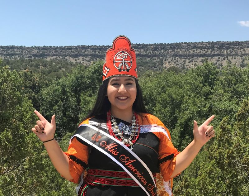 Miss American Indian OSU Cydnee pictured standing outside, wearing the MAIOSU crown and sash, along with traditional dress, smiling at the camera.