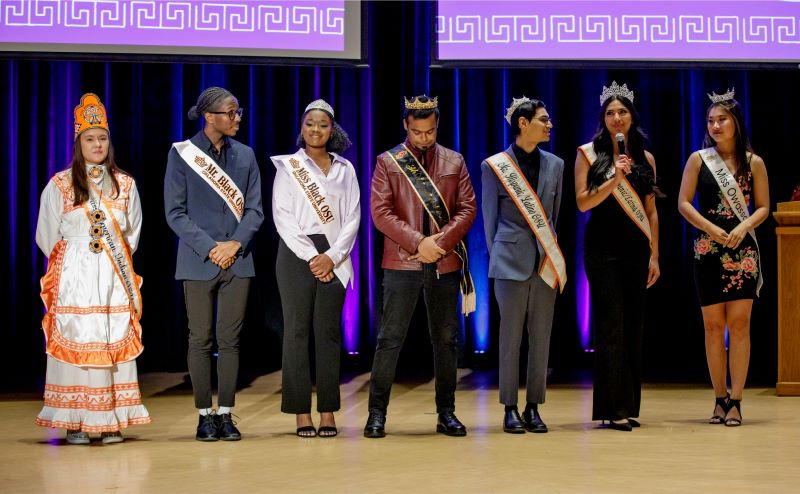 Miss American Indian OSU Shaeleigh, wearing an orange and white Choctaw dress along with her crown and sash, standing on stage with other OSU royalty members, all wearing their crowns and sashes.
