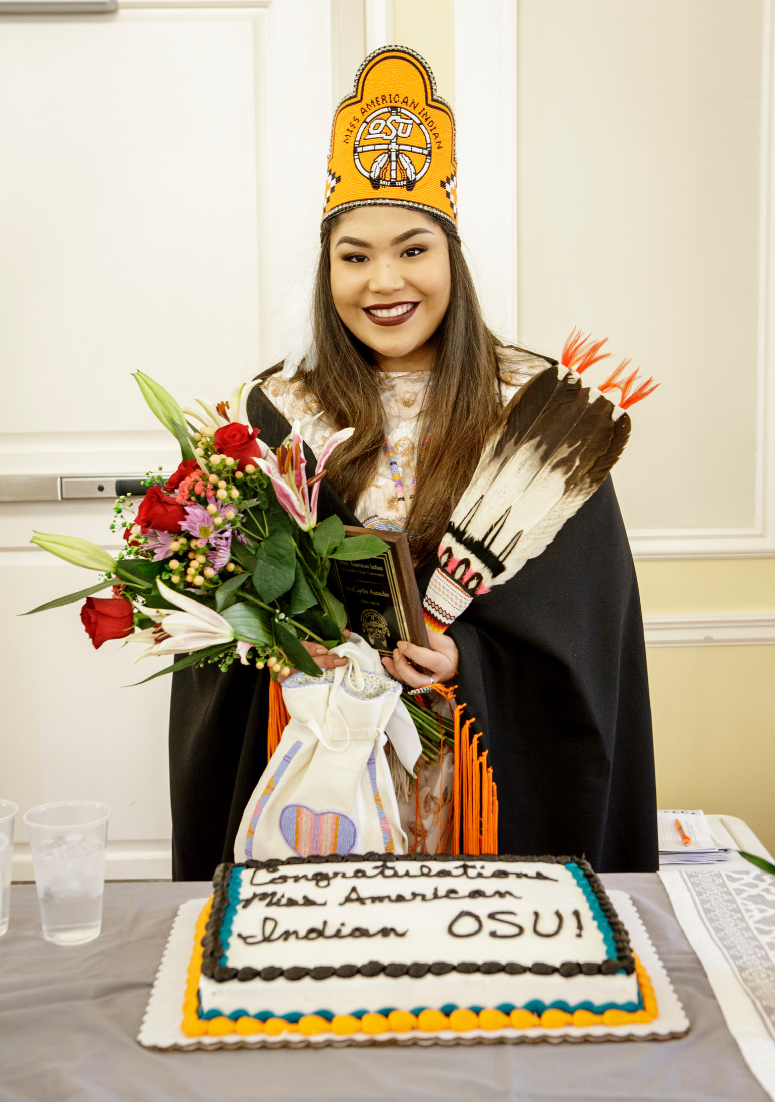 Miss American Indian OSU MaCaylin pictured smiling, wearing her traditional clothing, MAIOSU crown, shawl, and sash, standing with a cake labeled “Congratulations Miss American Indian OSU!”
