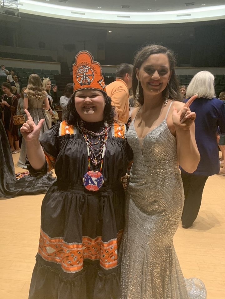 Miss American Indian OSU Kait pictured wearing Cherokee Tear Dressclothing and the MAIOSU crown, posing with Hadley, a contestant from the Miss Oklahoma State University pageant, who is wearing a silver ball gown.