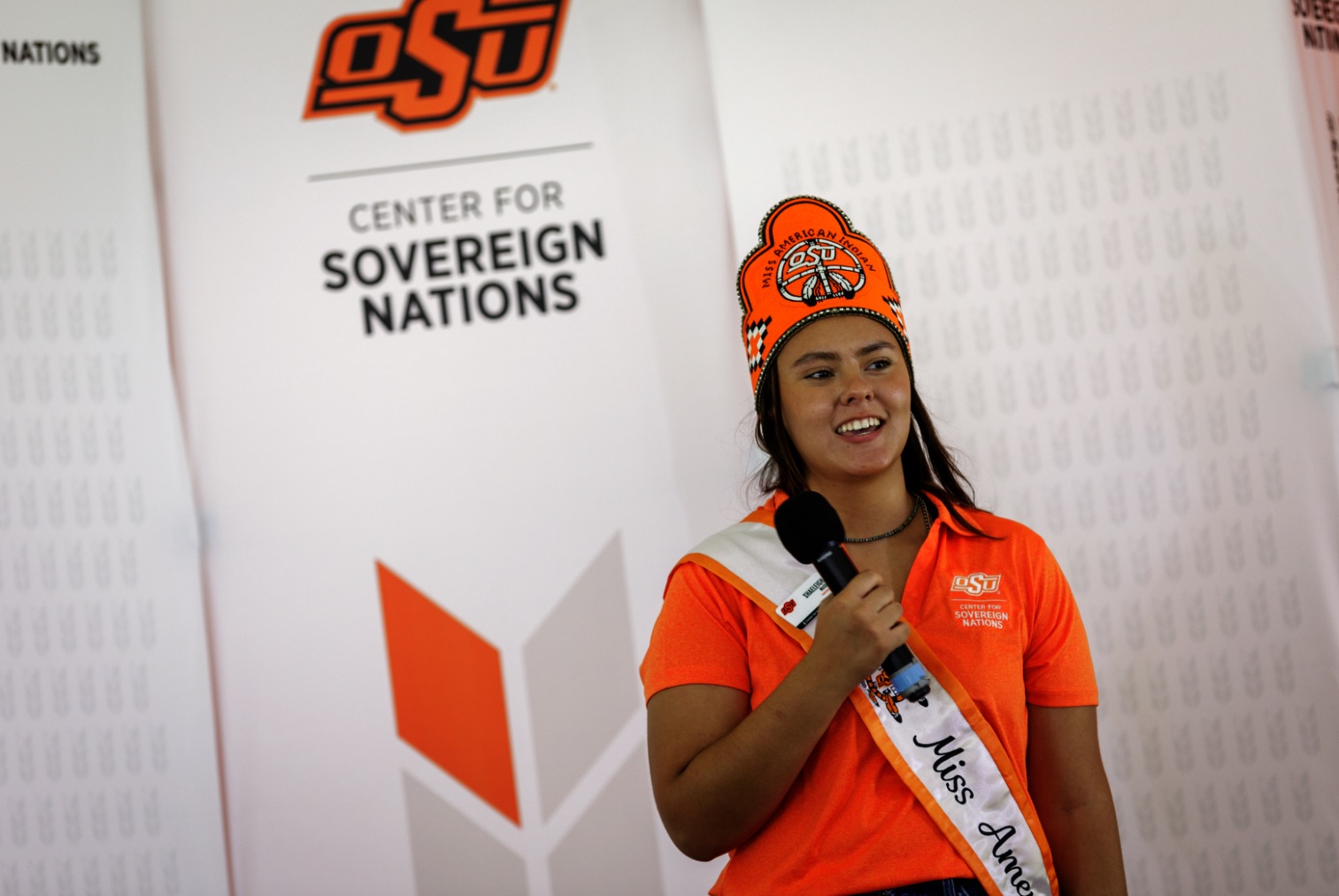 Miss American Indian OSU Shaeleigh, wearing the MAIOSU crown and sash along with an orange Center for Sovereign Nations polo, speaking into a microphone at an event.