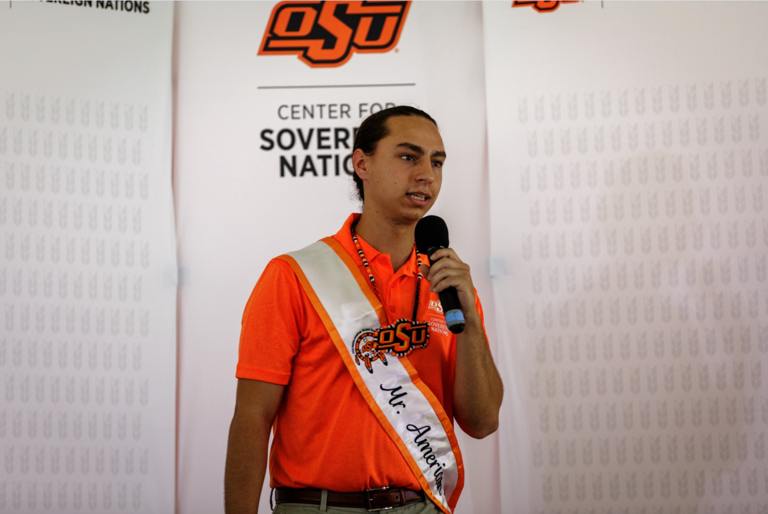 Mister American Indian OSU Caleb, wearing the MAIOSU sash and an OSU beaded medallion, along with an orange Center for Sovereign Nations polo, speaking into a microphone at an event.
