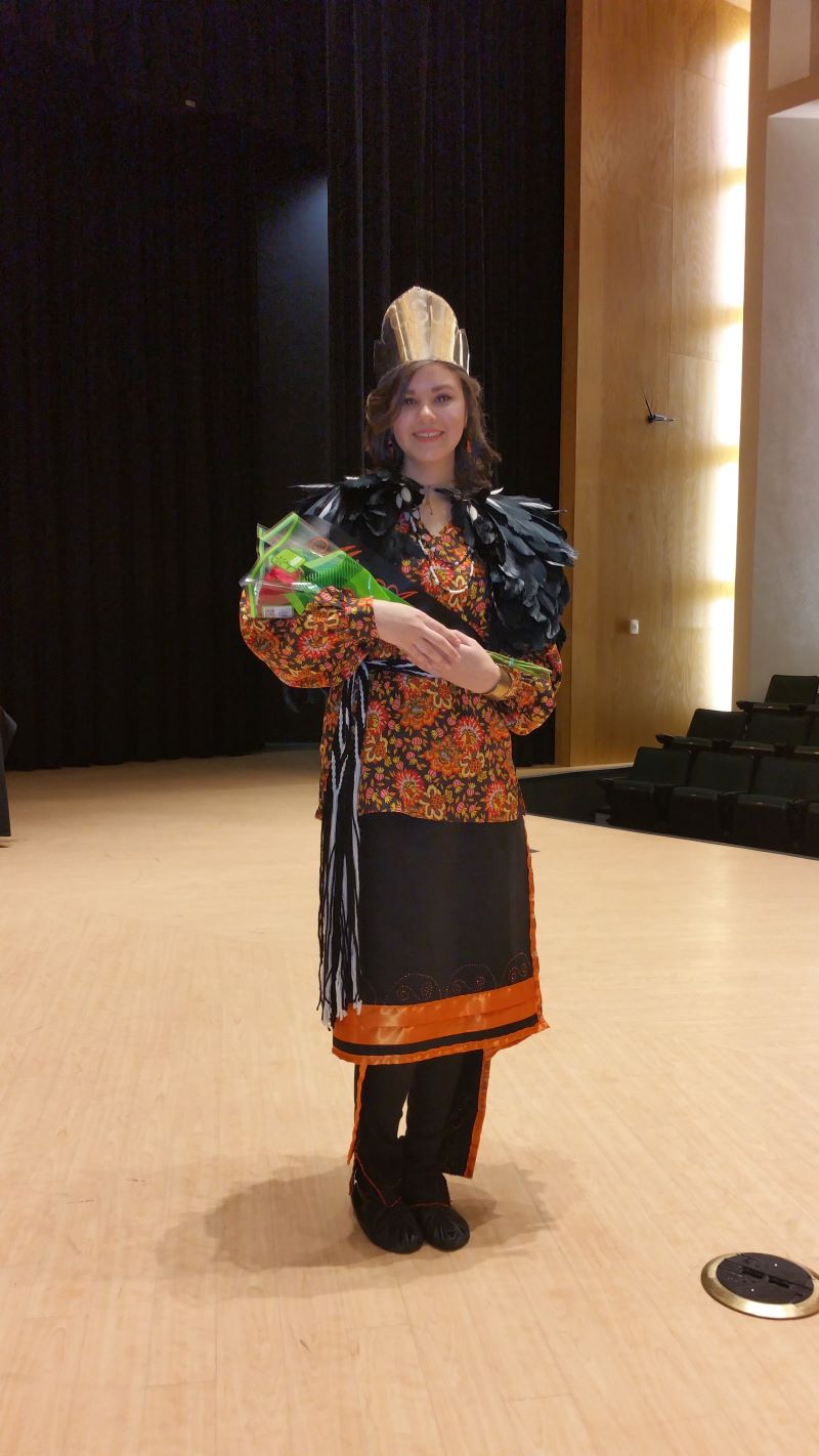 Miss American Indian OSU Meg pictured holding flowers after winning Miss American Indian OSU, wearing a Cherokee feather cape and Cherokee cloth blouse and skirt.