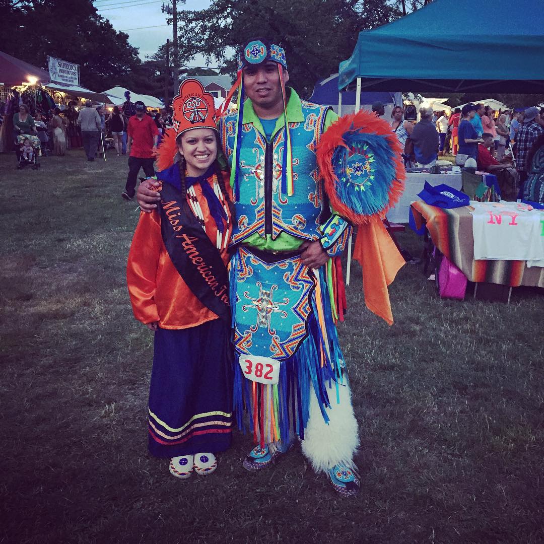 Miss American Indian OSU pictured wearing Southern Cloth regalia, posing with a male fancy dancer wearing traditional regalia at a powwow.