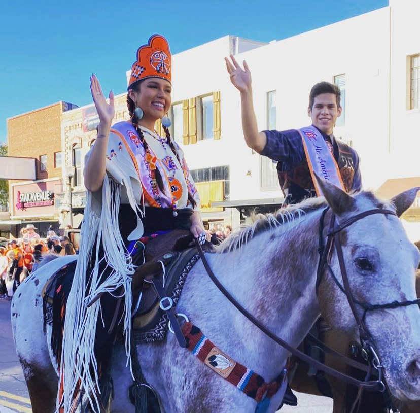 Miss American Indian OSU Gabby and Mister American Indian OSU Luke riding horses in the OSU Homecoming Parade, smiling and waving at the crowd.