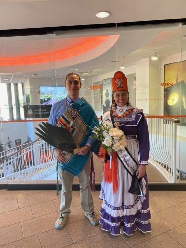 Miss American Indian OSU Shaeleigh, wearing a purple and white traditional Choctaw dress, and Mister American Indian OSU Caleb, wearing a button-up and tie, wrapped in a Pendleton blanket and holding a feathered fan, standing and smiling for the camera after winning the 2023–2024 MAIOSU pageant.