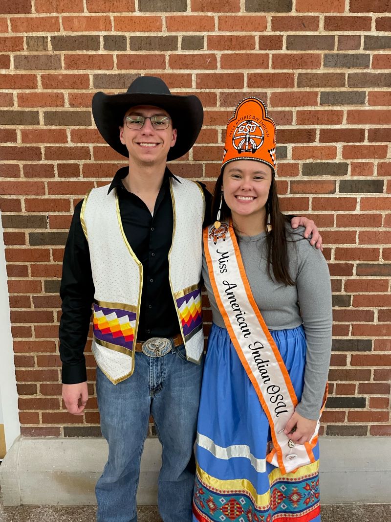 Miss American Indian OSU Shaeleigh and Mister American Indian OSU Caleb smiling together. Shaeleigh is wearing a ribbon skirt, crown, and sash, while Caleb is wearing a button-up shirt, and cowboy hat.