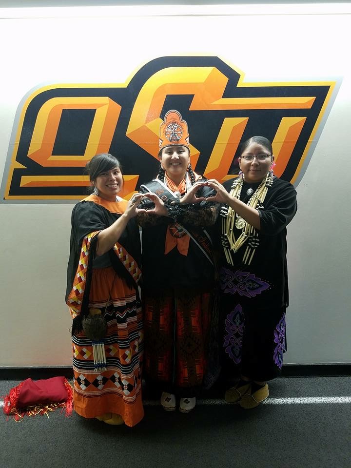 Miss American Indian OSU Cydnee pictured posing with two friends, all wearing women’s traditional regalia, in front of the OSU logo.