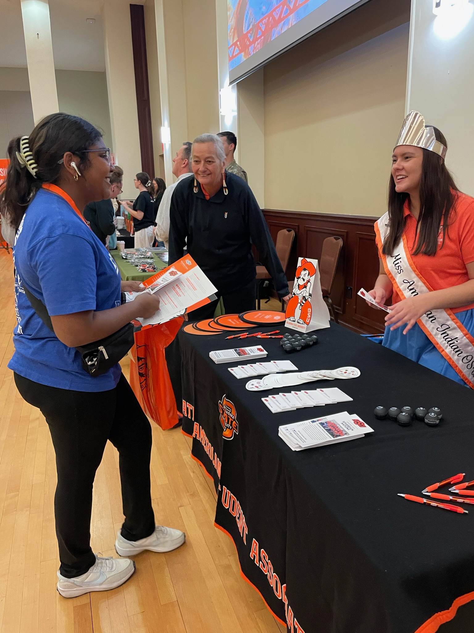 Miss American Indian OSU Shaeleigh, wearing a sash and ribbon skirt, and NASA co-advisor Dawna Riding-in-Hare at a tabling event, speaking to a guest.