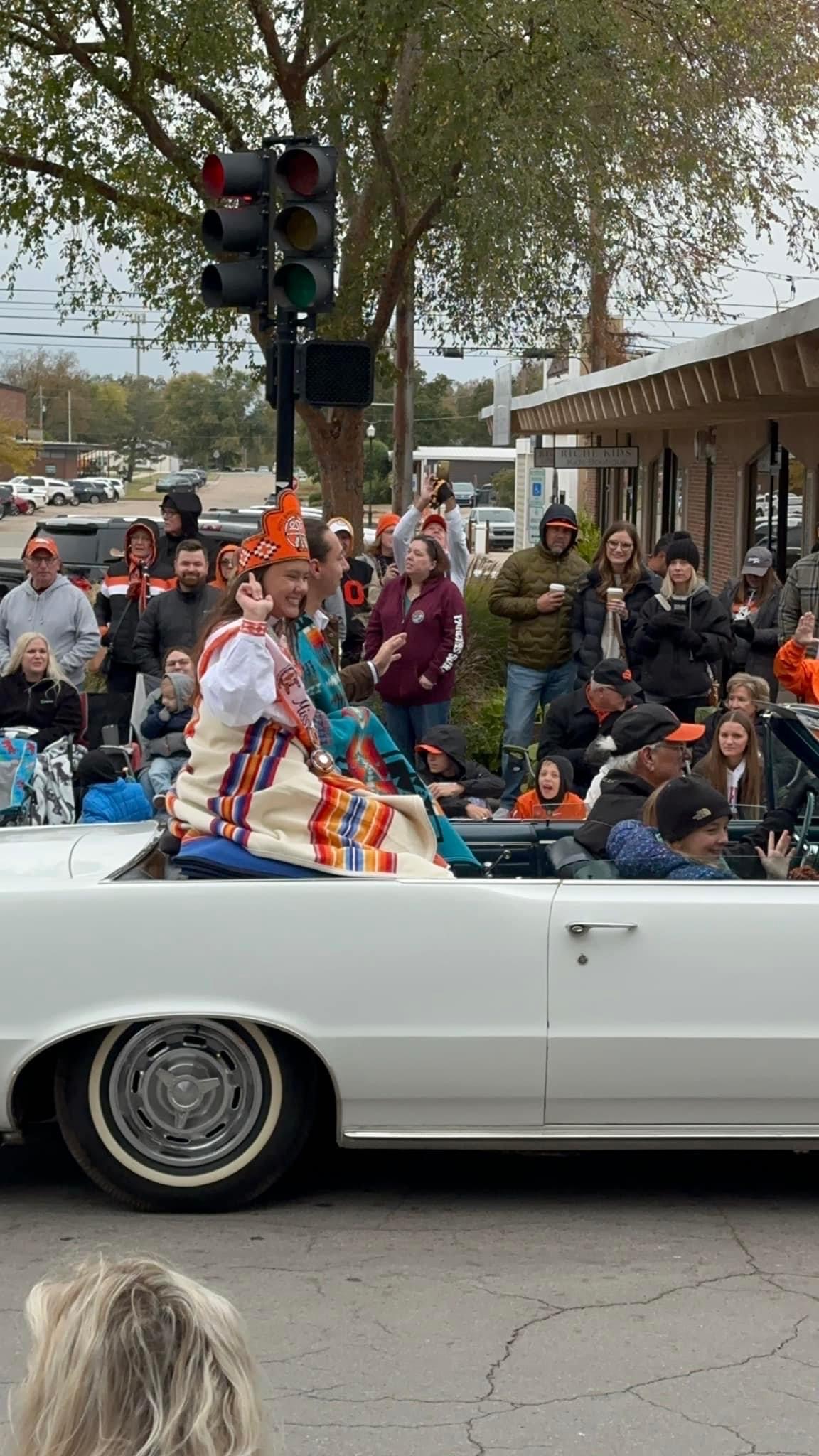 Miss American Indian OSU Shaeleigh and Mister American Indian OSU Caleb sitting in a convertible car, wrapped in Pendleton blankets, smiling and waving at the crowd at the OSU Homecoming Parade.