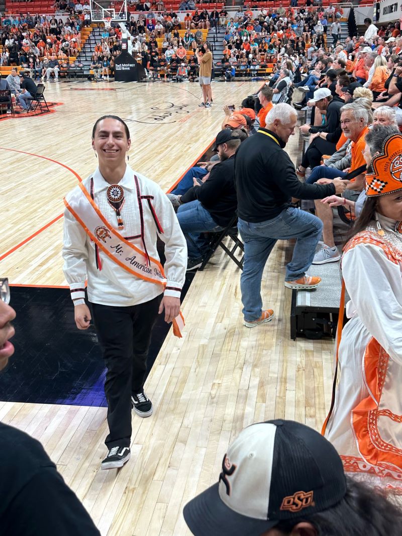 Mister American Indian OSU Caleb, wearing traditional clothing and a sash, standing on a basketball court and smiling at the N7 event.