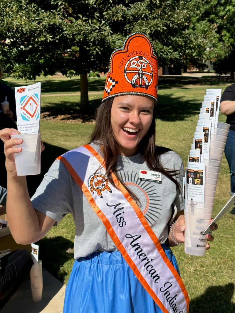 Miss American Indian OSU Shaeleigh pictured handing out flyers about Indigenous Peoples Day, wearing a ribbon skirt, sash and crown.