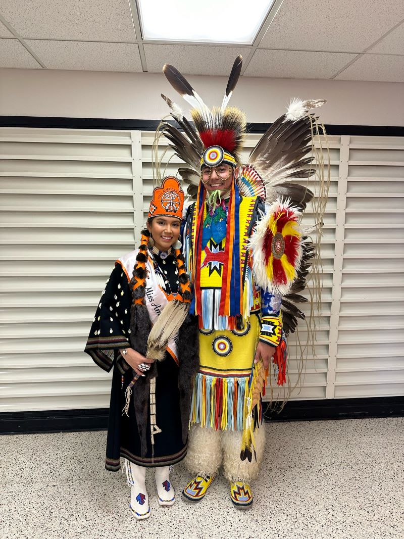 Miss American Indian OSU Gabby, wearing traditional regalia, posing for a photo with a fancy dancer in Fancy Dance regalia.