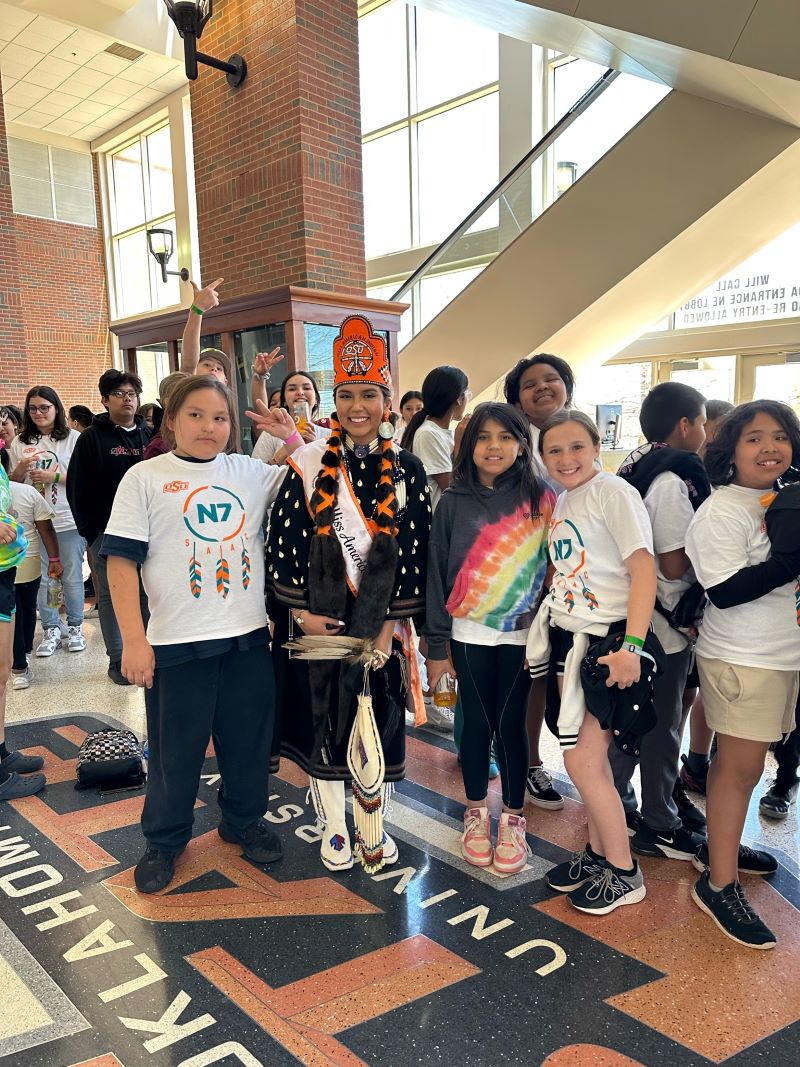 Miss American Indian OSU Gabby wearing crown and tradional clothing posing with children at N7 event Pictured.