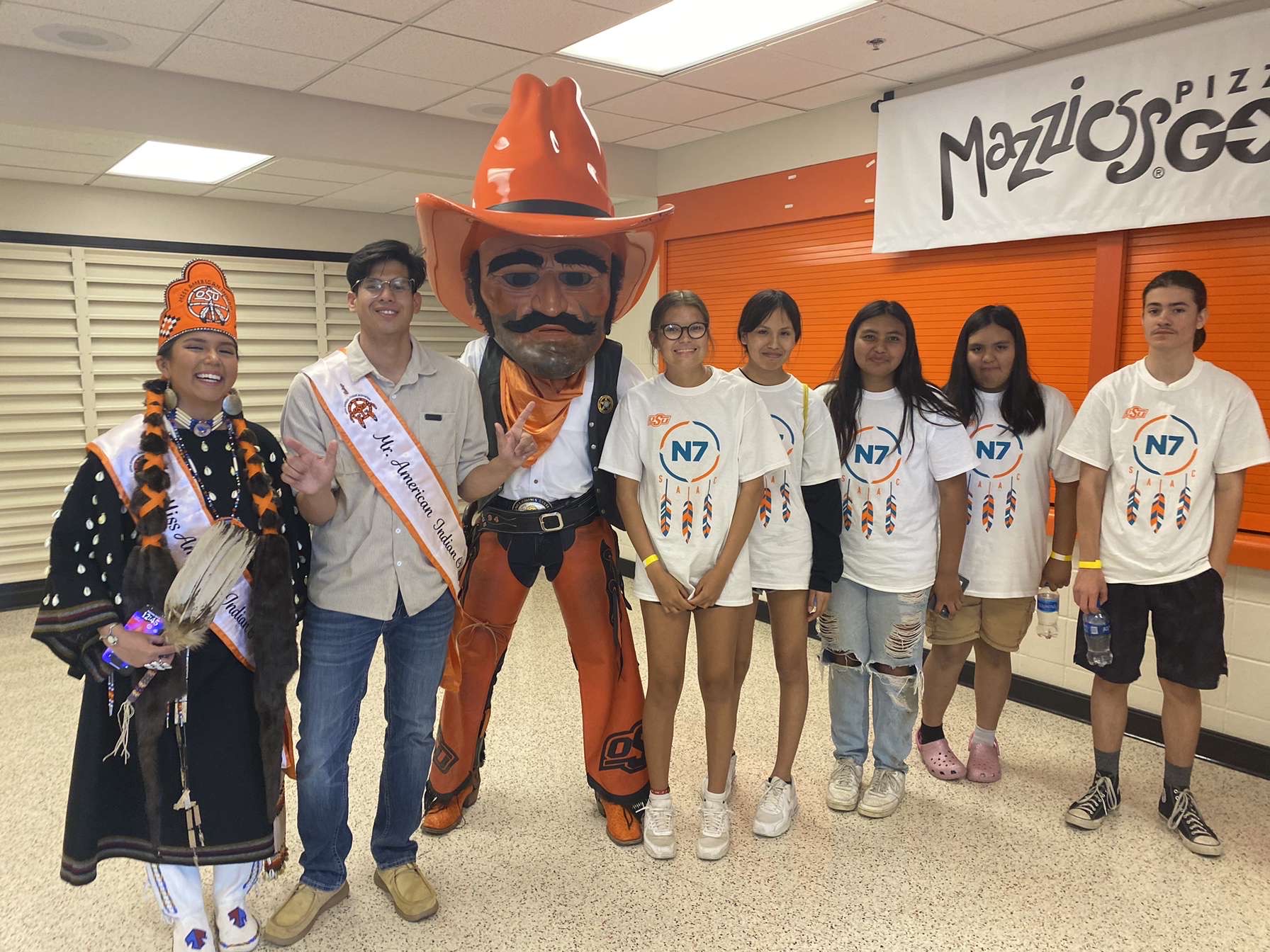 Miss American Indian OSU Gabby, wearing traditional clothing and holding a feather fan, and Mister American Indian OSU Luke, posing with Pistol Pete and an N7 child at the N7 event.