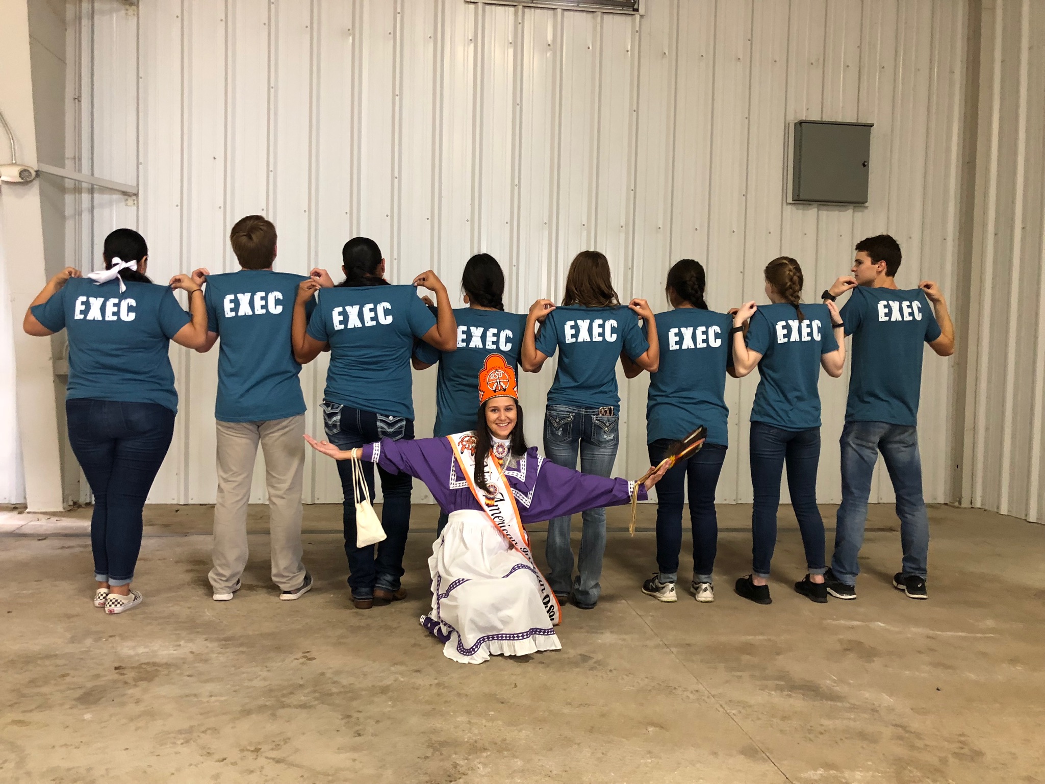 Miss American Indian OSU Cathrine, wearing a purple and white Choctaw traditional dress, smiling with the NASA executive team in a playful manner. Cathrine is crouched on the ground with arms open, smiling, while the executives are turned around, showing their matching shirts with “EXEC” written on the back.