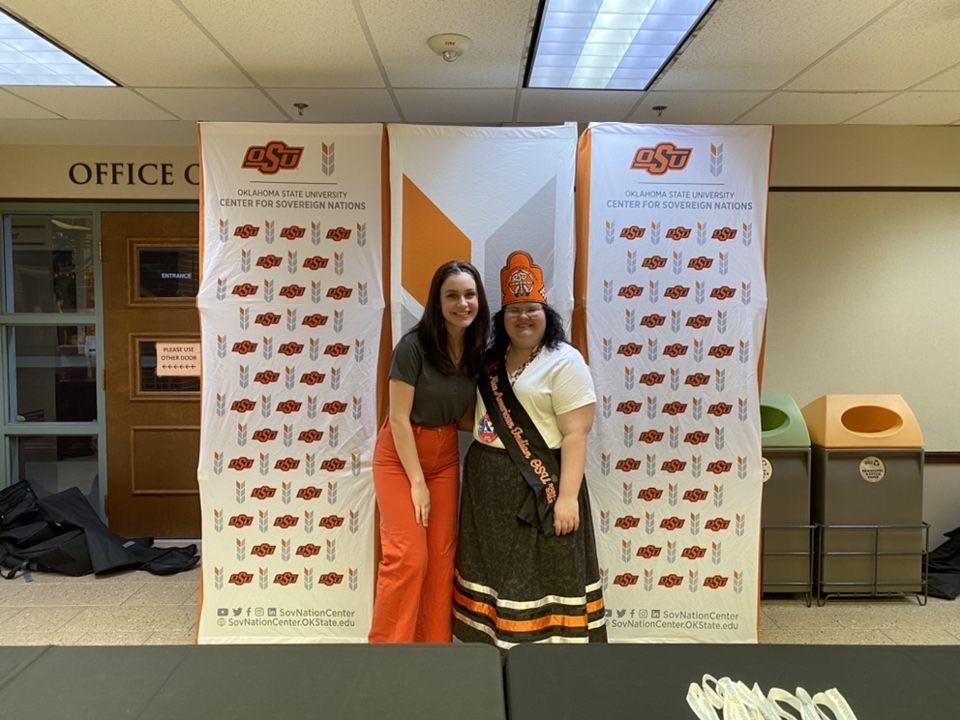 Miss American Indian OSU Kait pictured with her friend Tylie, smiling at the camera in front of Center for Sovereign Nations popup signs.