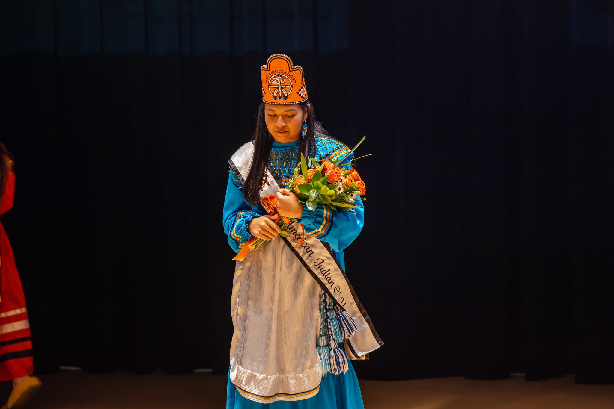 Cydnee Miller after being crowned Miss American Indian OSU 2025, holding an orange rose bouquet, and wearing the Miss American Indian OSU crown and sash.