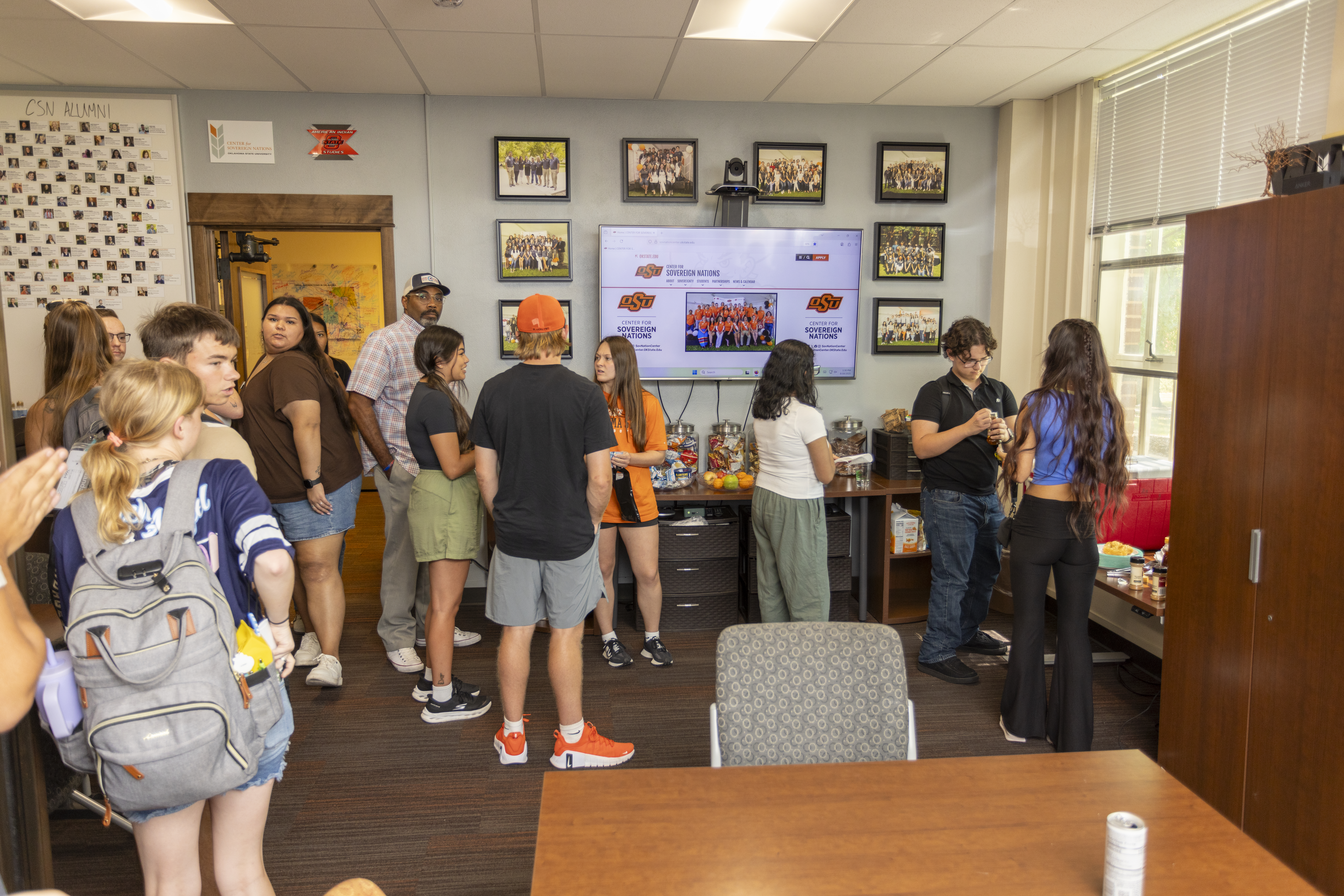 A group of people socializing in a room with a large monitor on the wall displaying a website and snacks on a table with frybread being served.