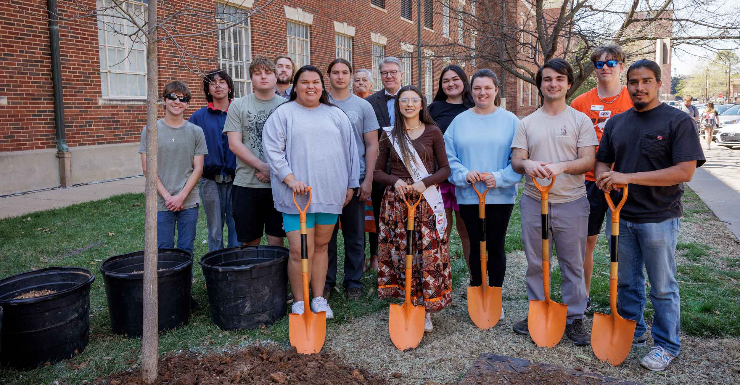Fourteen people smiling for the camera while holding orange shovels in front of a brick building