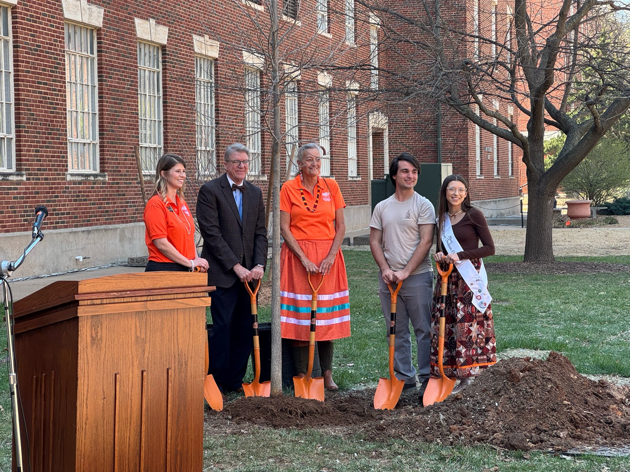 Five people holding shovels in front of a brick building smiling at the camera 