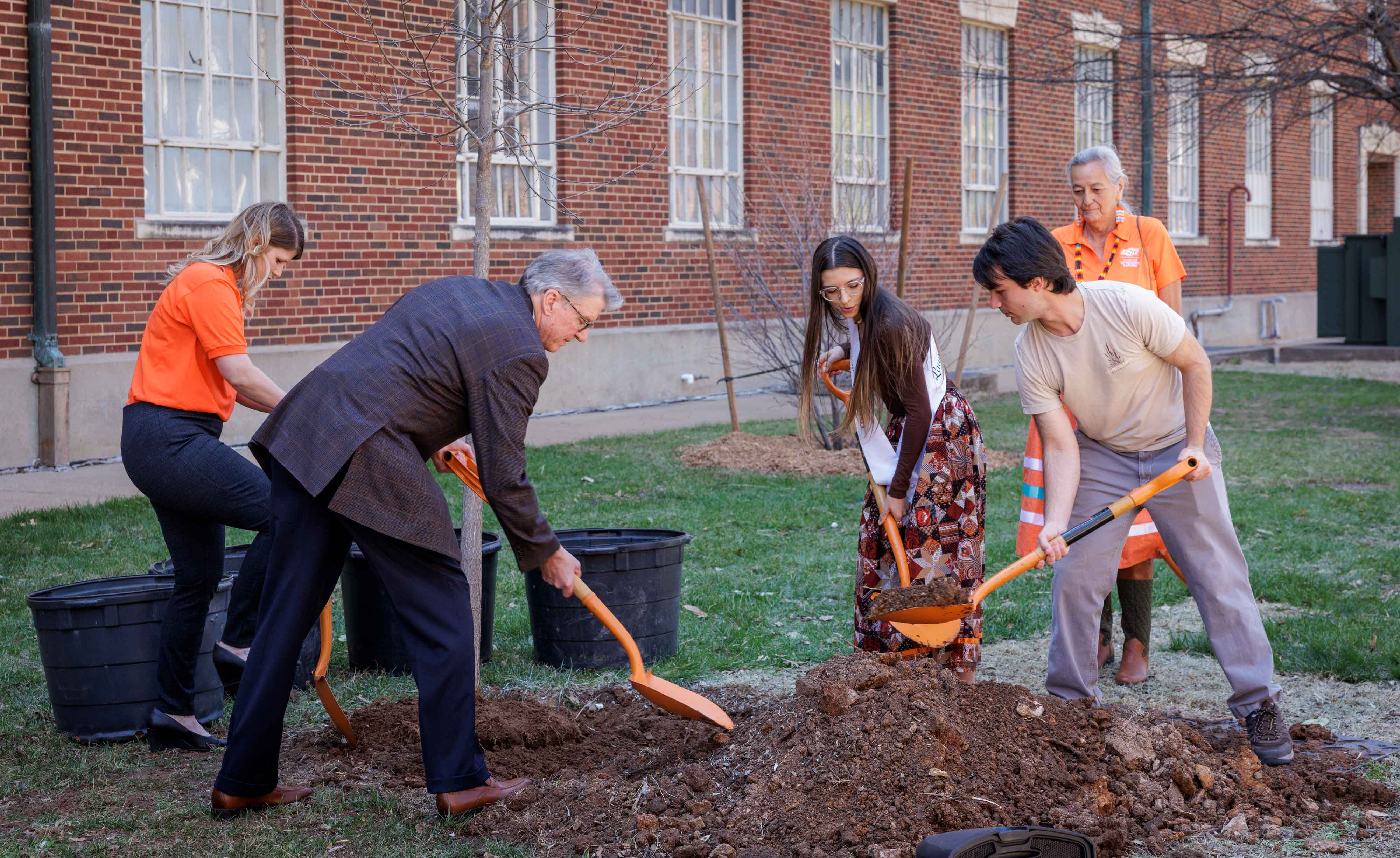 Group of four people planting a tree outside a brick building with orange shovels.
