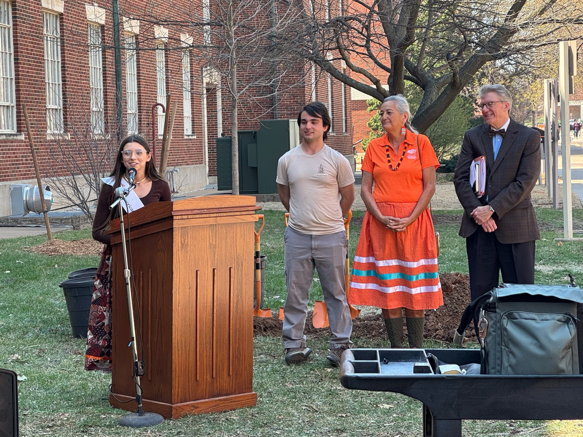 Miss American Indian OSU giving Lauren Branham giving a speech behind a podium outside a brick building with three people standing to the side.