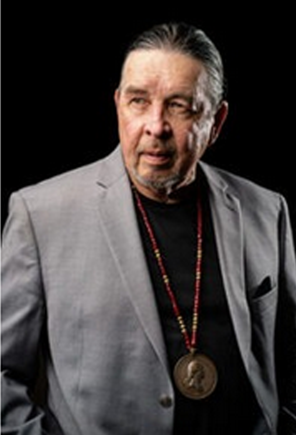 Harvey Pratt in a gray blazer with a medallion necklace on a black background.