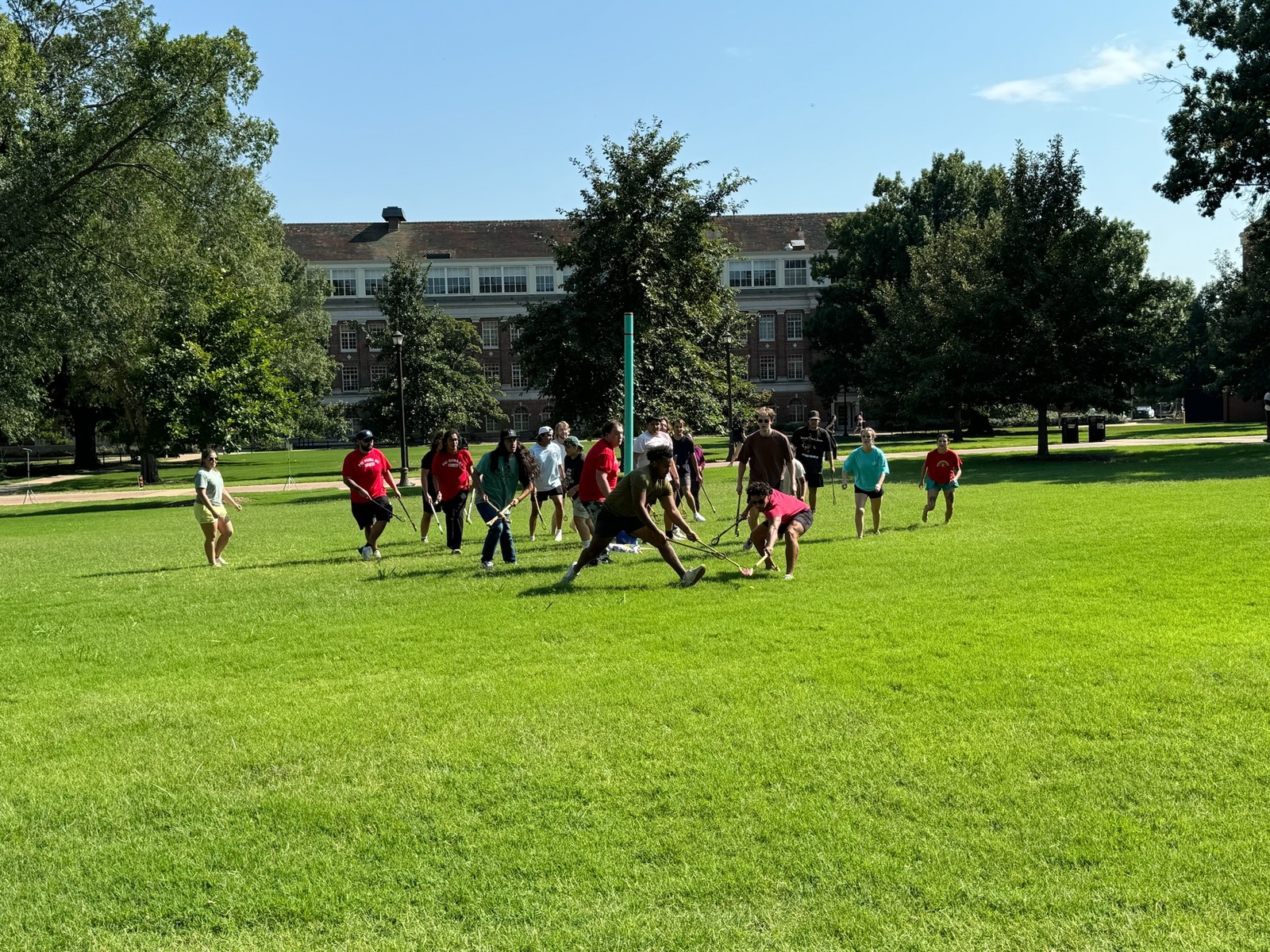 A group of people playing stickball on a grassy field with a brick building in the background.