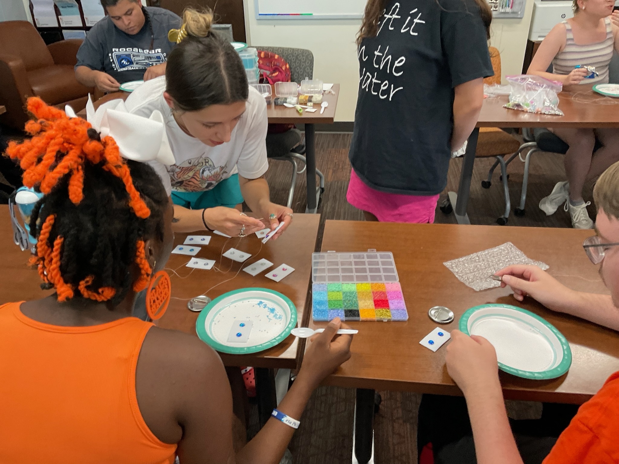 A person demonstrates beadwork to two others at a craft table with colorful beads and supplies.