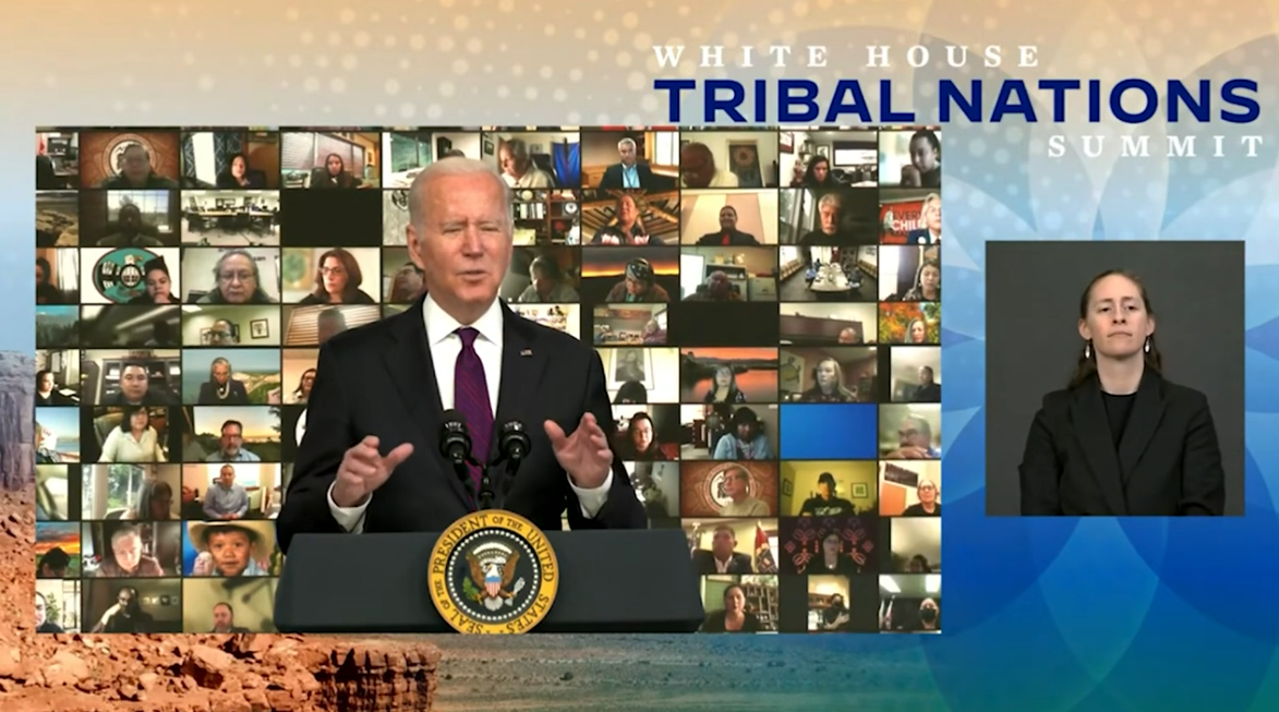 President Joe Biden a podium speaks against a backdrop of video call participants and a sign language interpreter, with a "White House Tribal Nations Summit" banner.